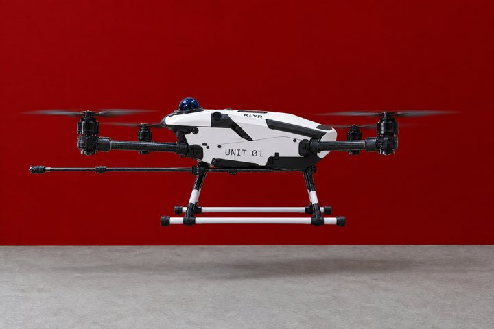 The white KLYR drone UNIT 01 hovering low in front of a red wall, its lance pointing forward under the fuselage.